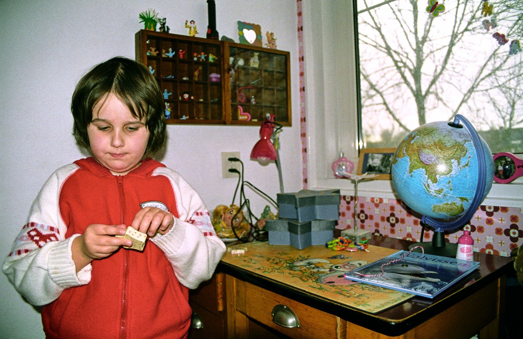 Our daughter in a red and white vest, standing next to a small wooden desk. It has drawers with metal handles. On the desk are blue boxes, a globe, a book about whales and a small light. Through the window tree branches without leaves.