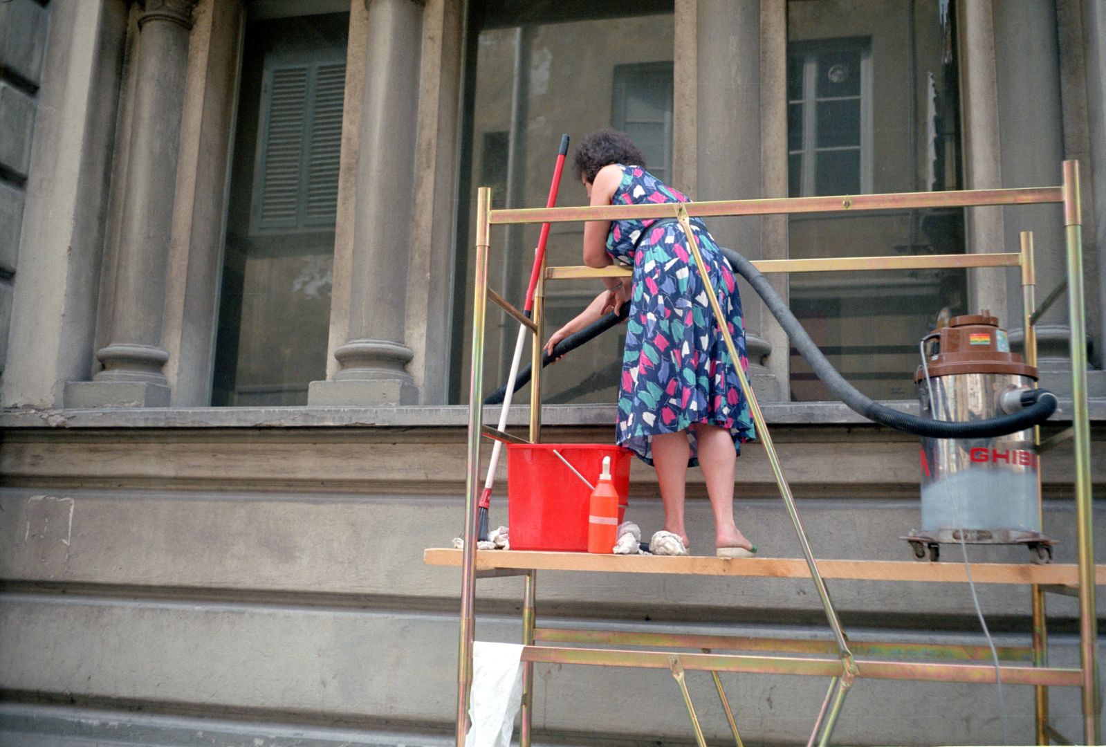 A woman is standing on scaffolding outside a building, cleaning a large stone windowsill with a vacuum cleaner. She is wearing a colorful dress with an abstract pattern, featuring shades of blue, pink, and green.

Next to her, a red bucket and a bottle are placed. The scaffolding itself is gold-colored.