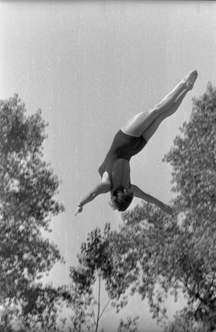 Swimmer jumping from high diving board, arms wide sidewards, head first, almost vertically. Picture is in black and white, and has scratches as the negative was damaged.
