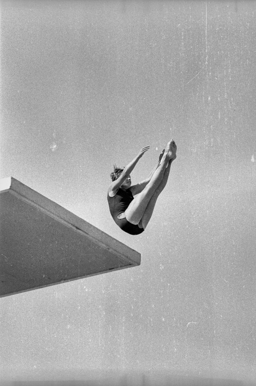 Swimmer jumping from high diving board, arms and legs pointed upwards. Picture is in black and white, and has scratches as the negative was damaged.