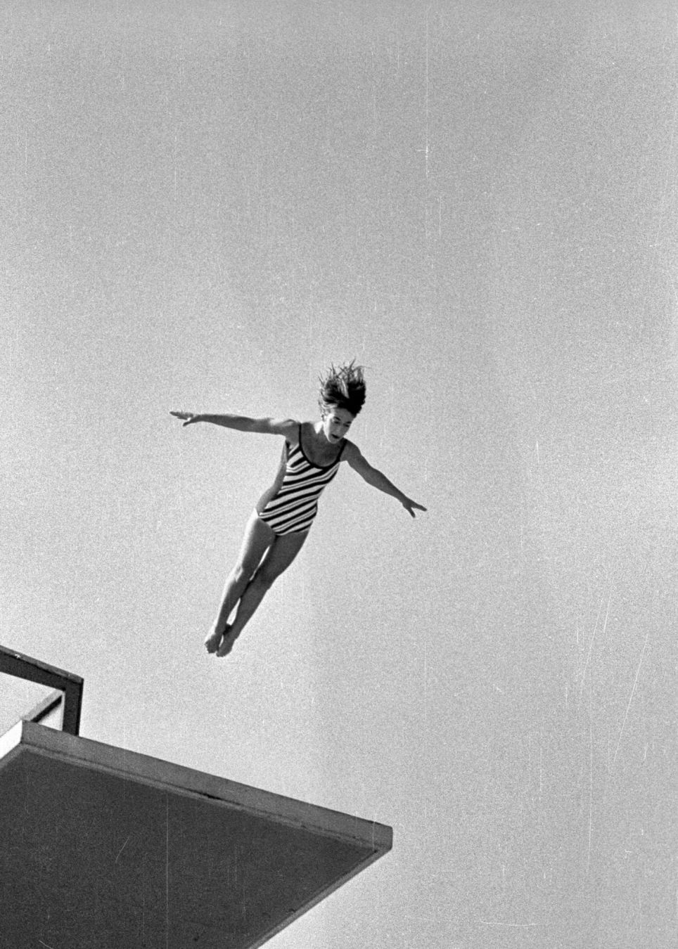 Swimmer jumping from high diving board, arms wide sidewards. Picture is in black and white, and has scratches as the negative was damaged.