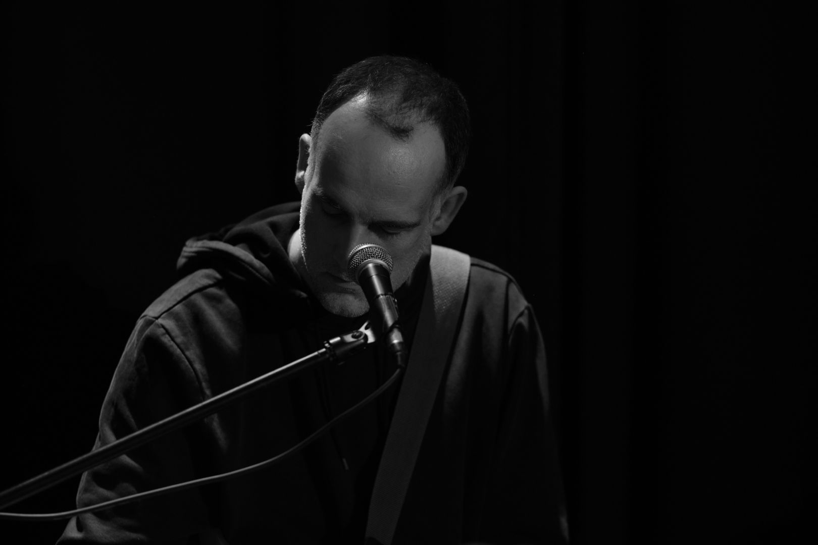 Man playing piano and guitar, mostly visible for the light on the contours of his body. His partly bald head has a great reflection. Picture has a completely black background, and is black and white.